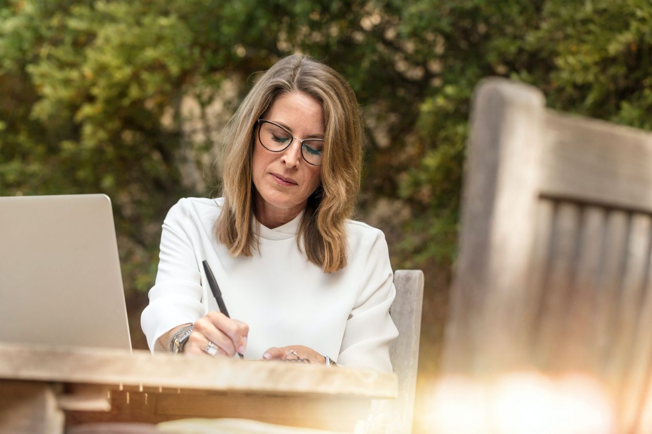 Older women sitting outside with her computer while she writes notes with a pen